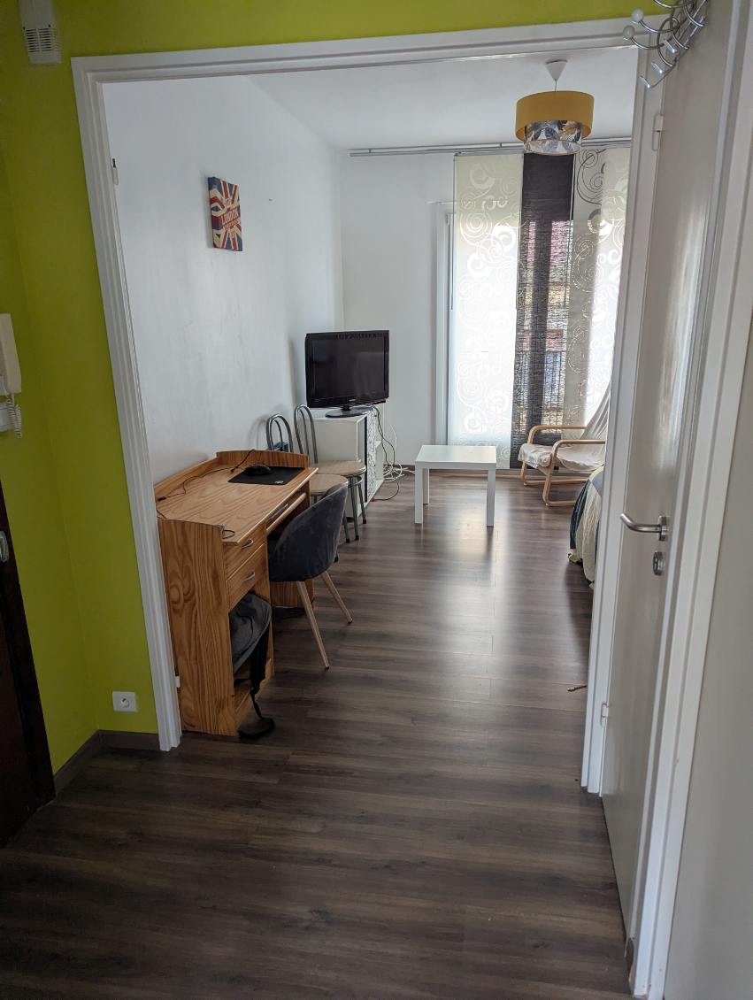 A view into the living space is presented, featuring a light wood desk with two chairs and a small white table. A television is positioned on a unit, while natural light filters through sheer curtains. The floor is finished in a dark, warm tone.