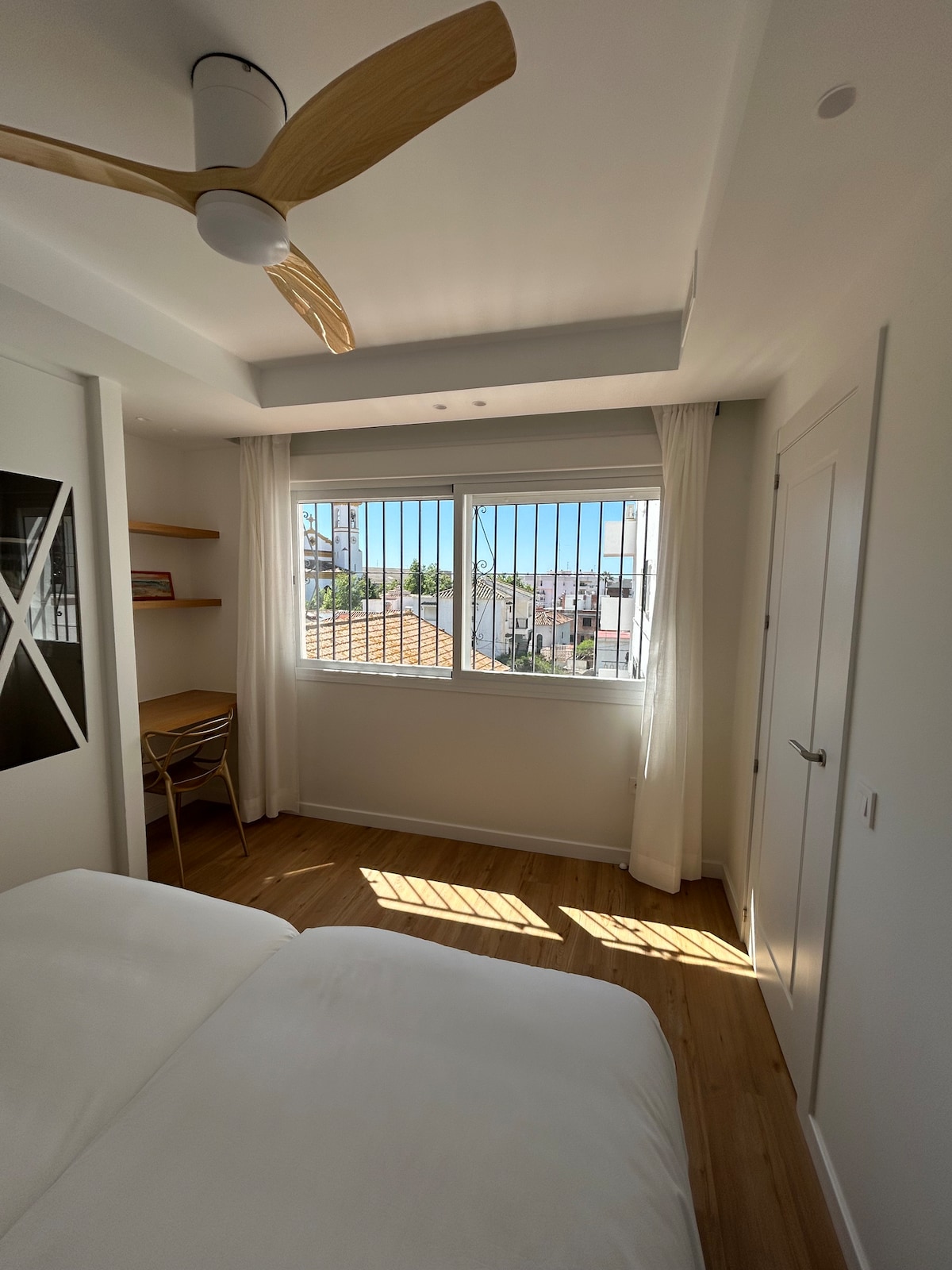 A bright bedroom features a spacious bed with crisp white linens. Natural light enters through a large window with sheer curtains. A wooden desk with a chair is positioned nearby, complemented by a shelf against the wall. A ceiling fan is mounted above.