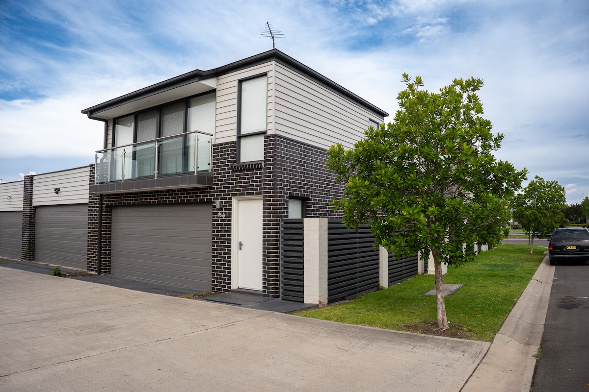 A modern building features a minimalist design with a blend of dark brick and light paneling. A private entrance is visible, along with a garage and a small landscaped area with greenery. The exterior is well-maintained, reflecting the contemporary style of the apartment.