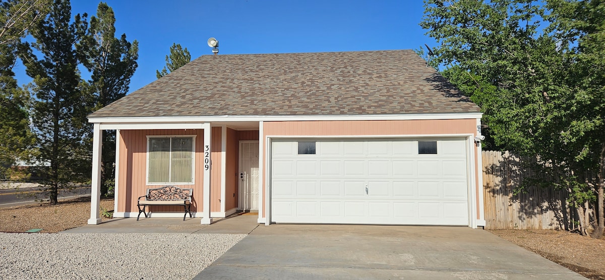 A single-story home with a light pink exterior is presented, featuring a gabled roof and a welcoming porch. A decorative bench is positioned by the front door, and a driveway leads to a white garage door. Lush trees are visible in the surroundings.