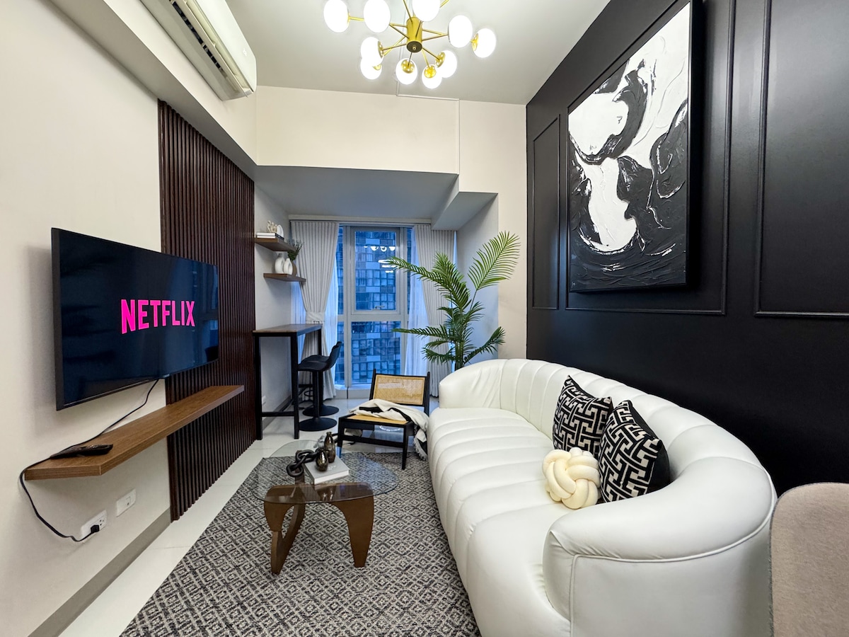 A bright living area features a white curved sofa and a dark accent wall holding a large abstract artwork. A wooden coffee table sits in front, while a television displays the Netflix logo. A vibrant plant adds a touch of greenery beside a window offering city views.