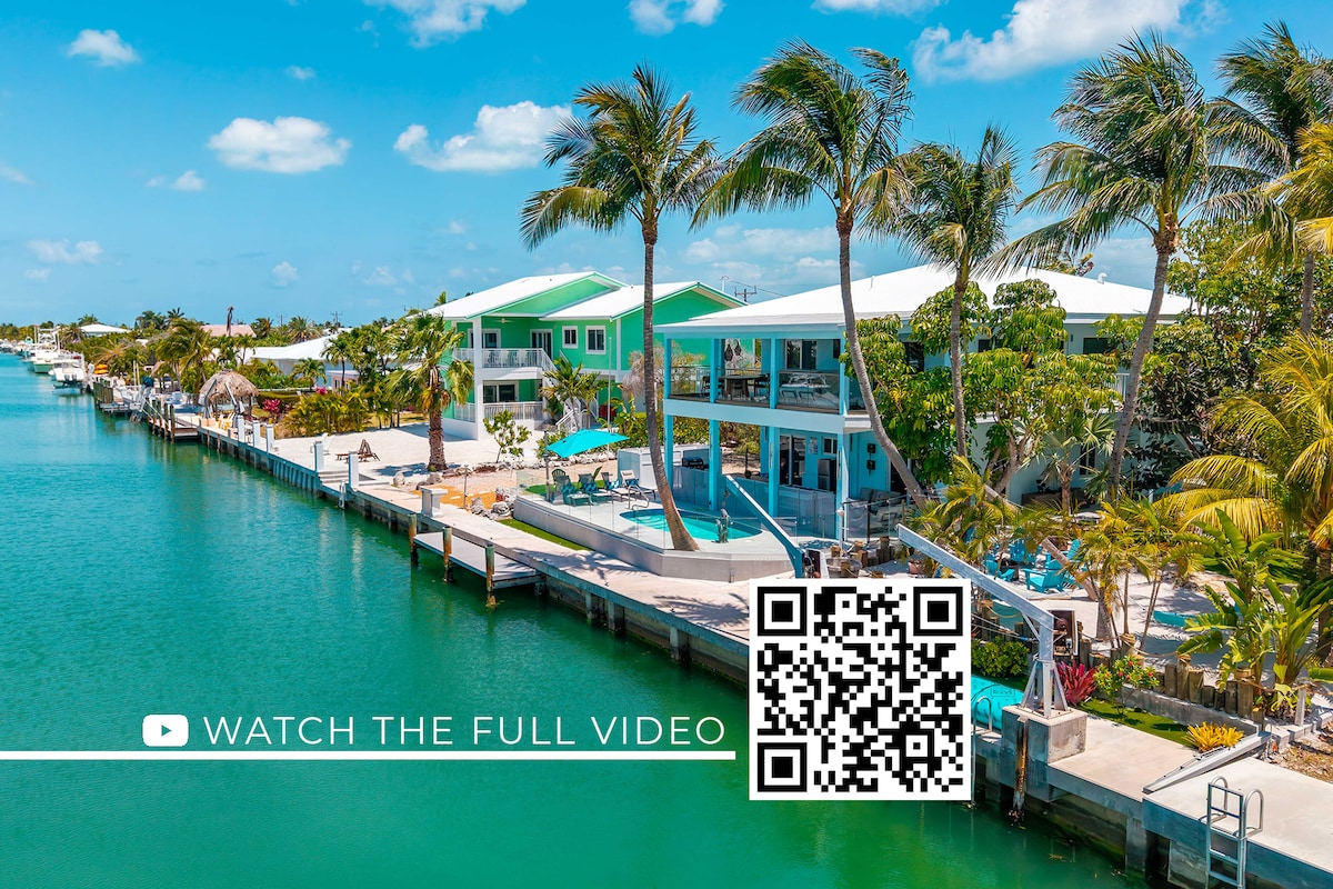 An aerial view captures the waterfront property with a heated pool and outdoor seating areas, surrounded by lush palm trees. The private canal is visible, along with neighboring homes. Blue skies enhance the tropical setting, creating a serene atmosphere perfect for relaxation.