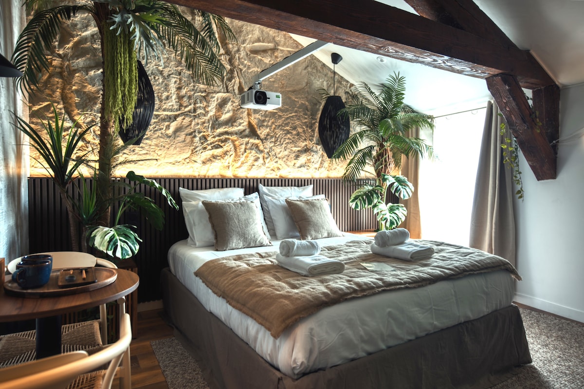A spacious room features a large bed adorned with soft pillows and a neutral-colored blanket. Surrounding the bed, lush greenery and decorative plants create a vibrant environment. A cozy seating area with a small table is visible, alongside a projector mounted on the ceiling projecting onto the wall.