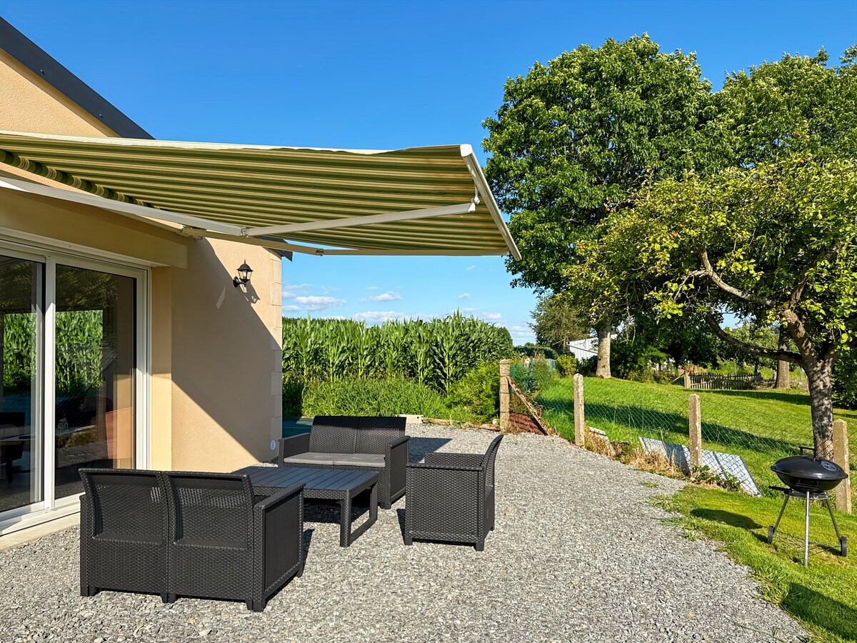 An outdoor seating area is featured under a retractable awning, with four dark wicker chairs surrounding a low coffee table. A barbecue grill is positioned nearby, with green grass and a tree in the background, adding to the relaxing outdoor atmosphere.
