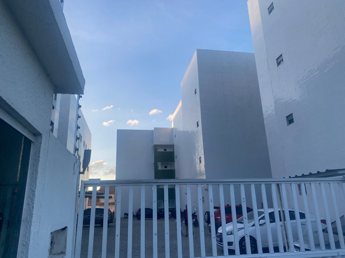 A view of the surrounding buildings is captured, featuring modern, white facades that reflect the sky. A paved parking area is visible in the foreground, with several parked cars. The image is taken from a gated entrance, overlooking the outdoor space in a well-maintained environment.