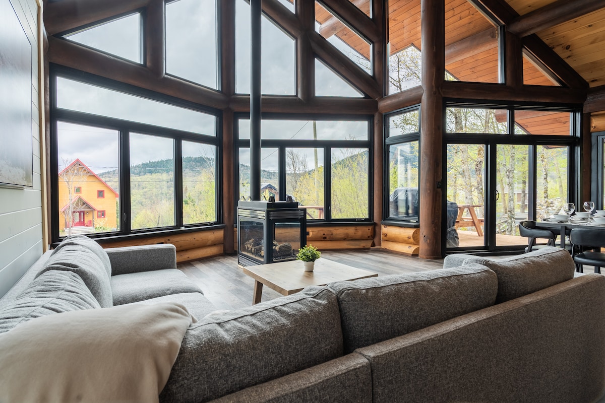 A spacious seating area is highlighted by large windows that allow natural light to fill the space. The room features a comfortable sectional sofa facing a modern coffee table, with a view of the surrounding landscape visible through the glass. A fireplace adds a cozy touch.