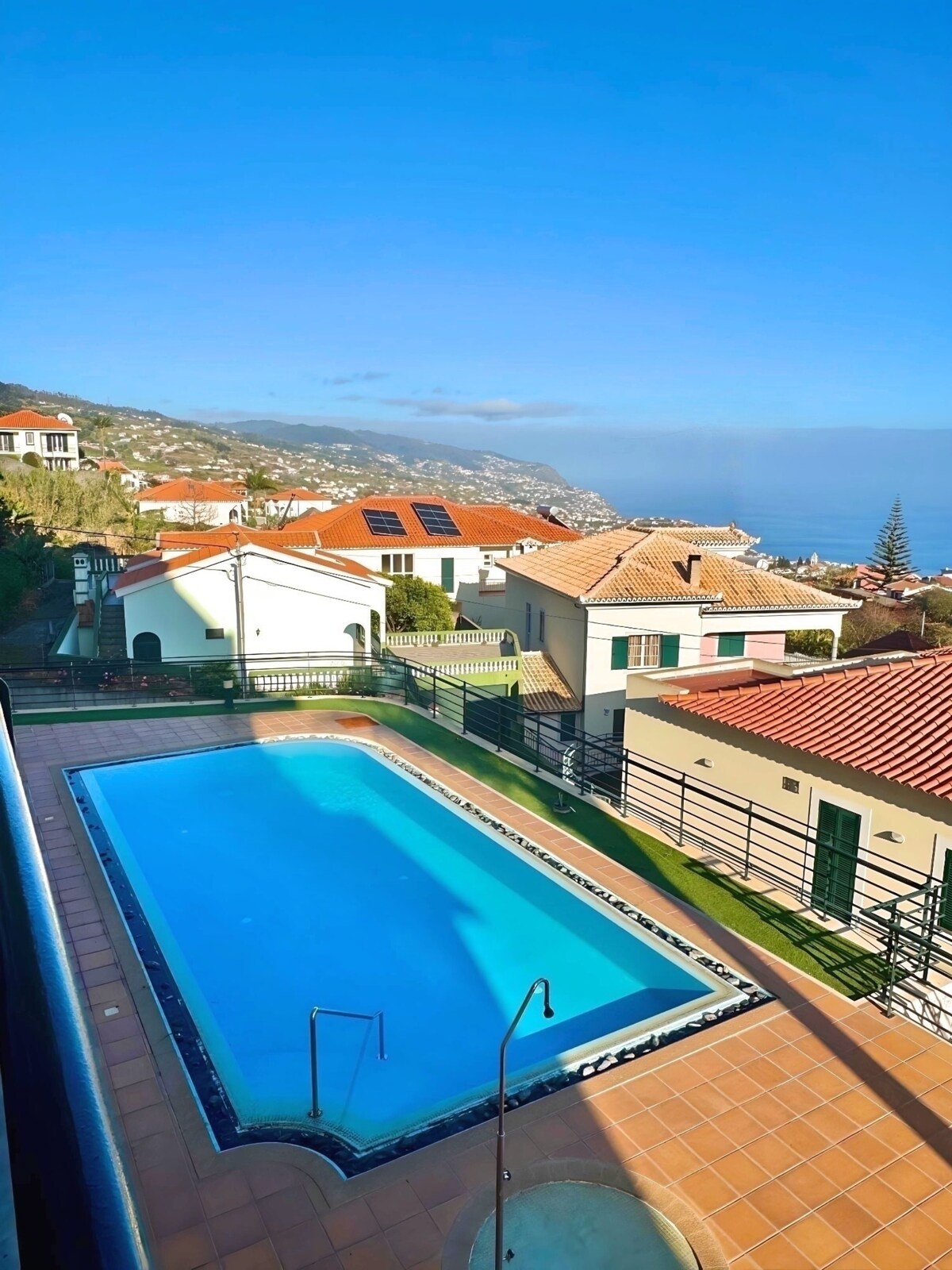 An aerial view captures the inviting swimming pool set within a landscaped area, surrounded by a fence. The azure water reflects the bright sky above, while the neighboring houses display a mix of red rooftops and light-colored facades, contributing to a serene residential ambiance.
