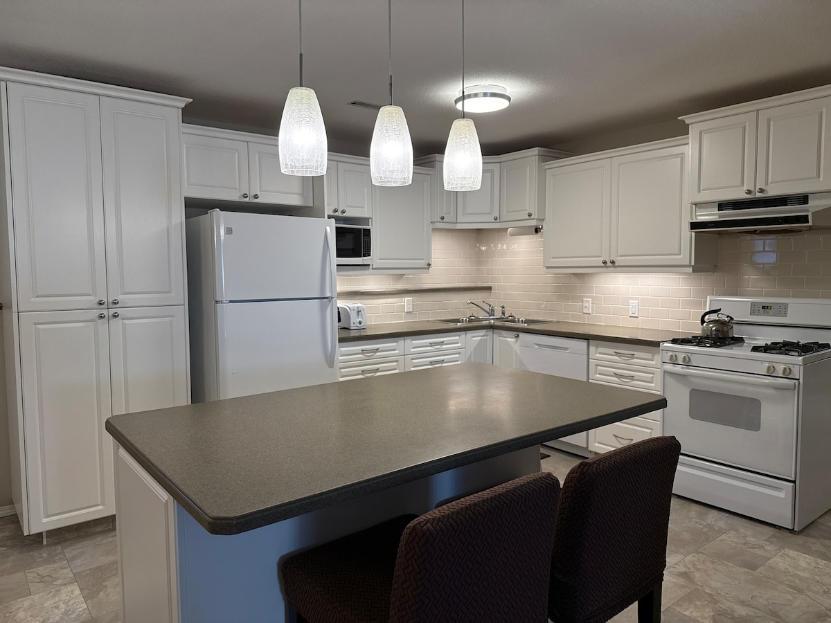 A modern kitchen is equipped with white cabinetry and stainless steel appliances. A refrigerator, microwave, and gas stove are visible. A central island with seating for two is featured, and pendant lights provide illumination. Beige tiles cover the floor, complementing the color scheme.