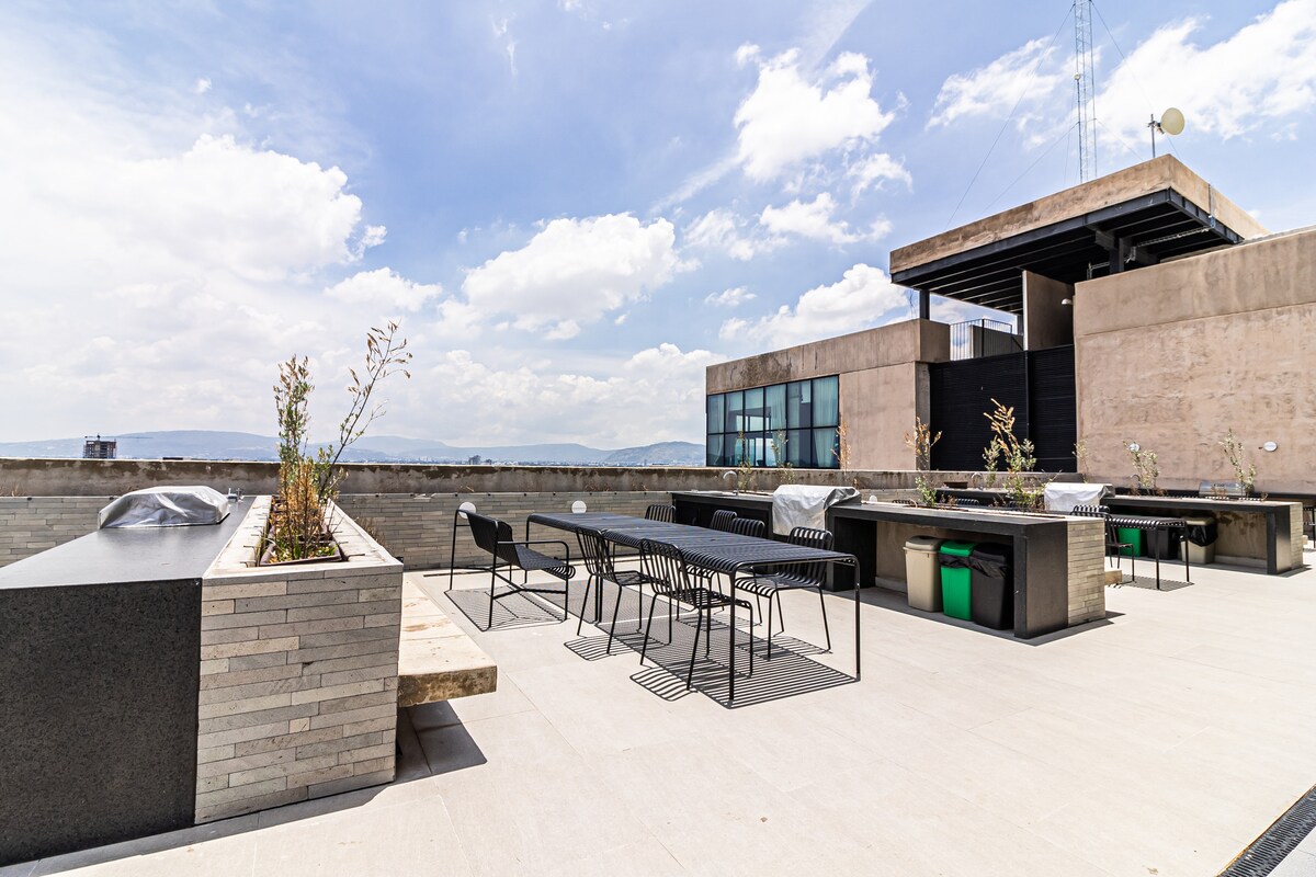 A spacious rooftop terrace is shown, featuring modern outdoor furniture including a long dining table and black chairs. Grassy planters line the perimeter, enhancing the open space that offers views of the surrounding area. The sky above is illuminated with scattered clouds.
