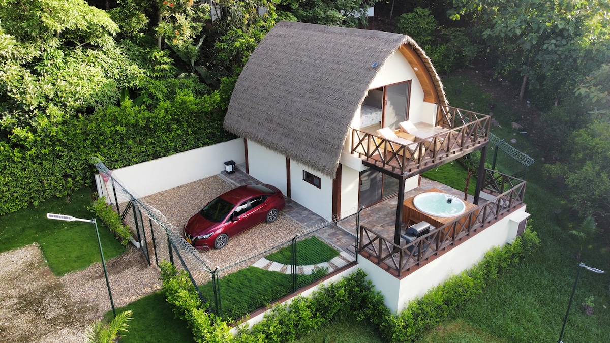 An aerial view of a two-story private cabin features a thatched roof and a spacious outdoor area. A red car is parked in a designated space surrounded by gravel. A private hot tub is visible on the balcony, alongside wooden railings offering a view of the lush greenery.