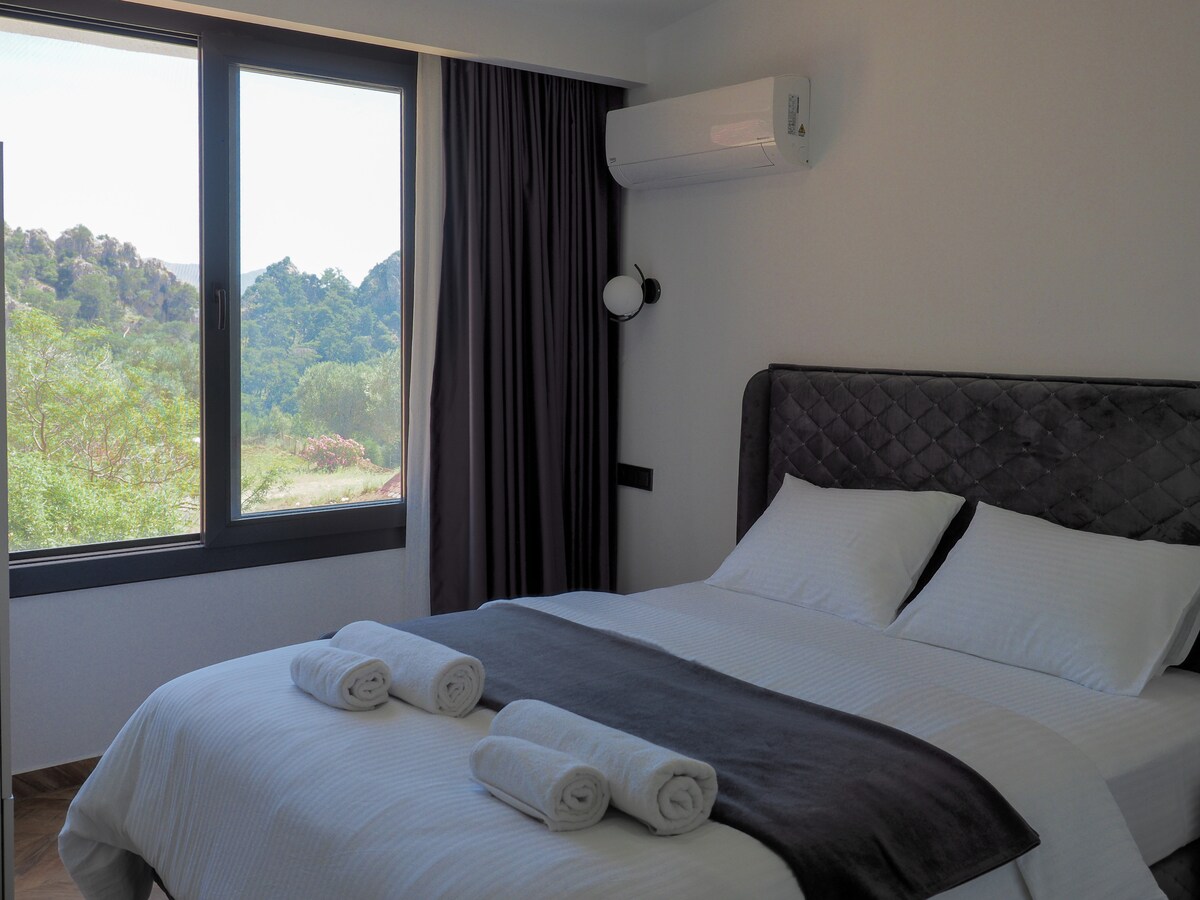 A modern bedroom features a spacious bed with crisp white linens and rolled towels placed neatly on the duvet. Large windows allow natural light to fill the room, offering views of green hills and the surrounding landscape.