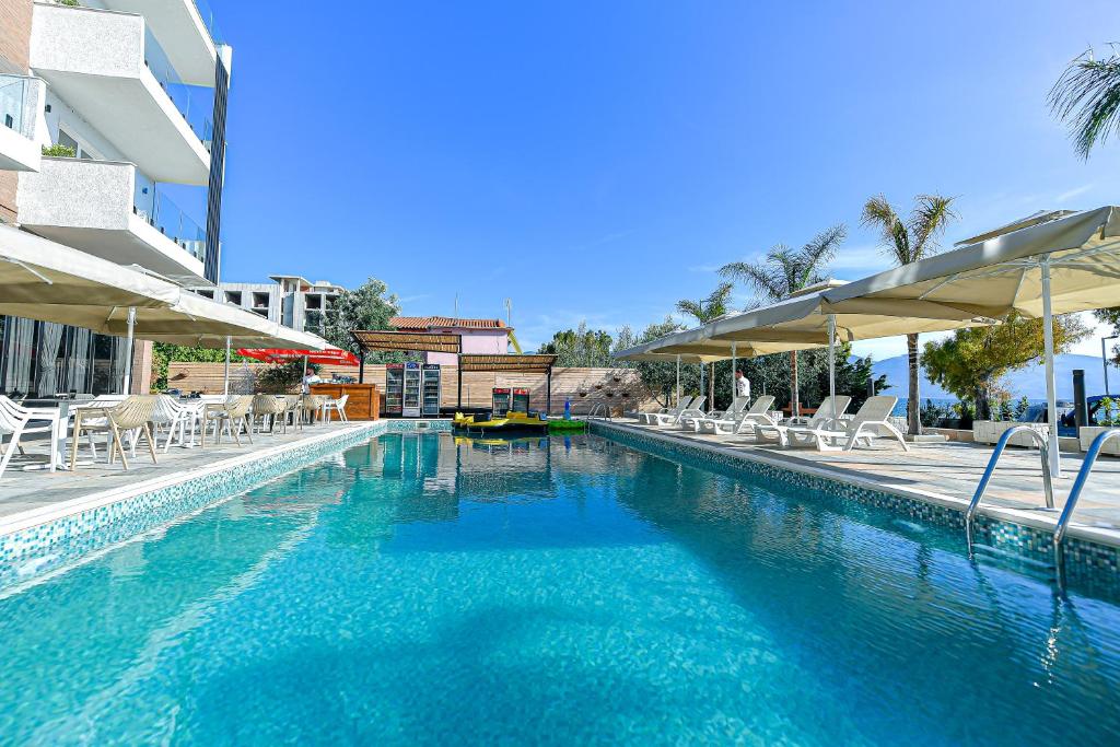 A clear outdoor swimming pool is surrounded by lounge chairs and shaded areas. Palm trees and buildings can be seen in the background against a bright blue sky. The pool's shimmering surface reflects natural light, inviting relaxation and enjoyment.