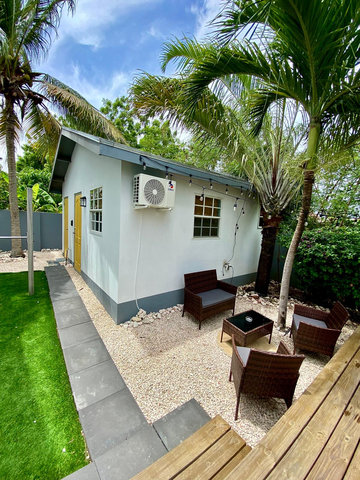 An outdoor seating area is visible, featuring two wicker chairs and a glass-topped table. The studio is framed by palm trees and situated within a landscaped yard, with a patch of green grass and stone pathways. An air conditioning unit is mounted on the exterior wall.