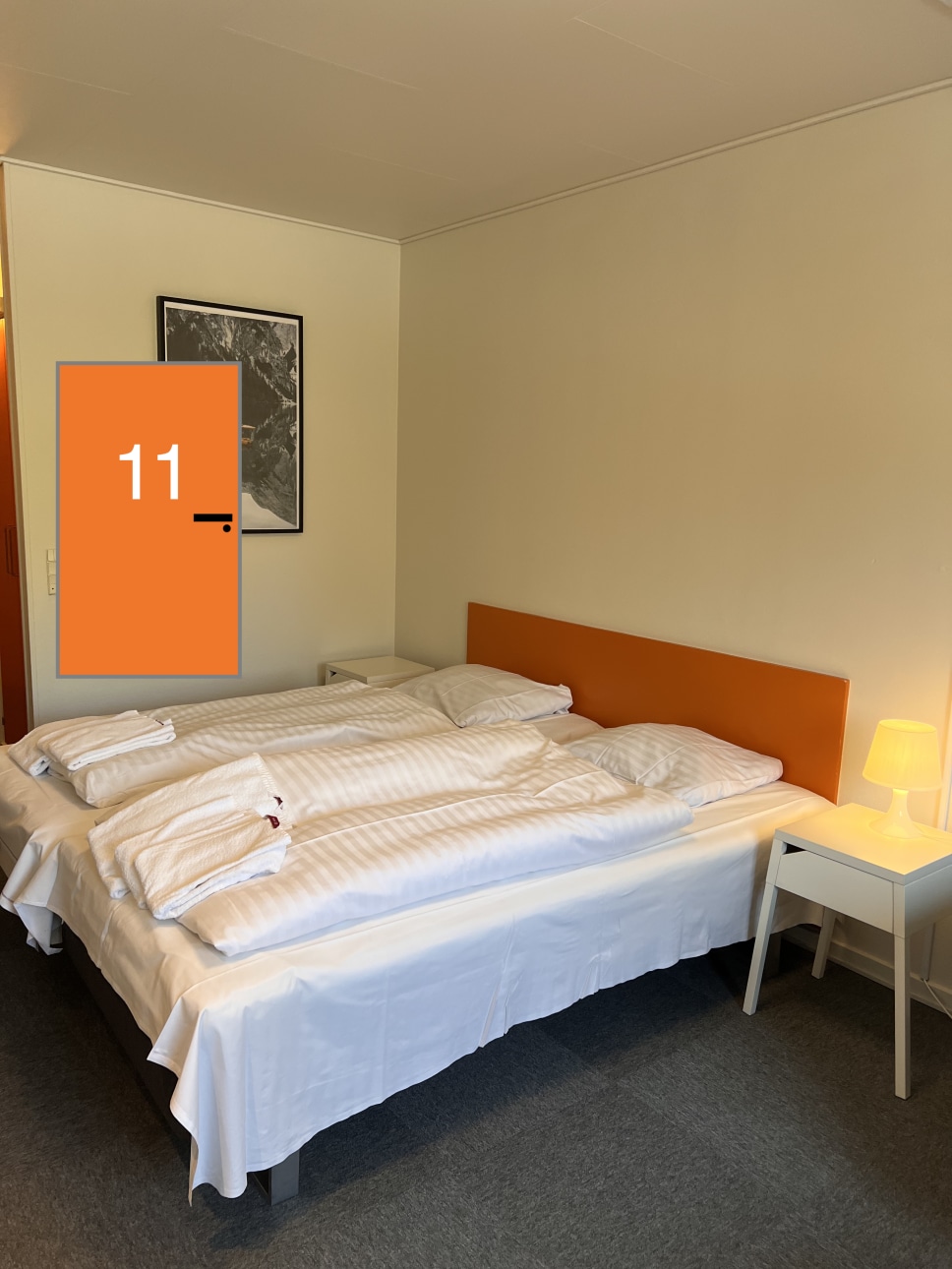 An inviting sleeping area is presented with a spacious bed covered in white striped linens. Two soft pillows and towels are neatly arranged on top. A small nightstand with a lamp accompanies the bed, while a simple artwork adorns the wall. The door to the room displays the number 11.