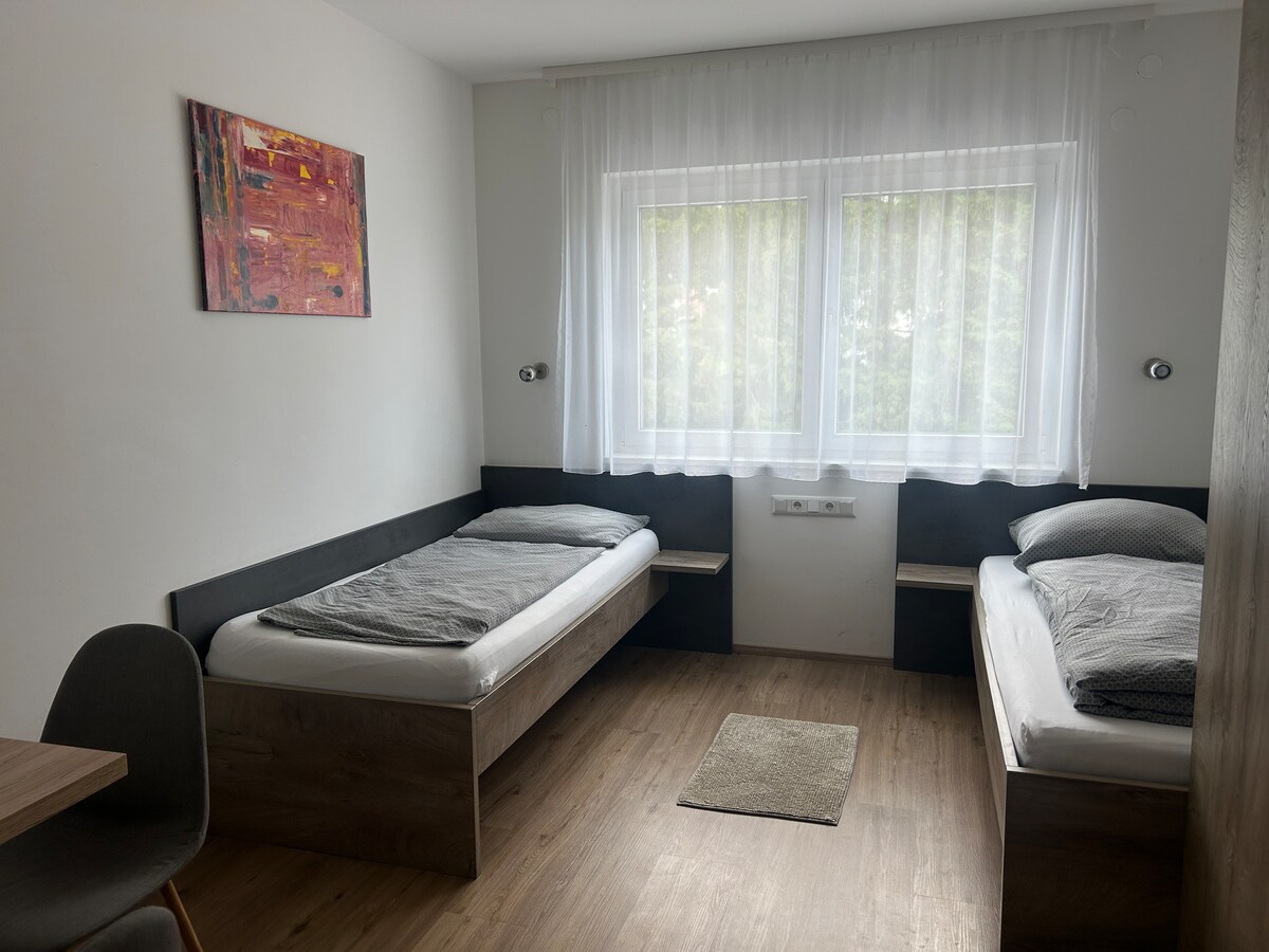 A simple and modern bedroom displays two spacious beds with gray linens, positioned on opposite sides of the room. A small rug sits on the wooden floor, and a table with a chair is visible nearby. Natural light flows through a large window dressed with white curtains.
