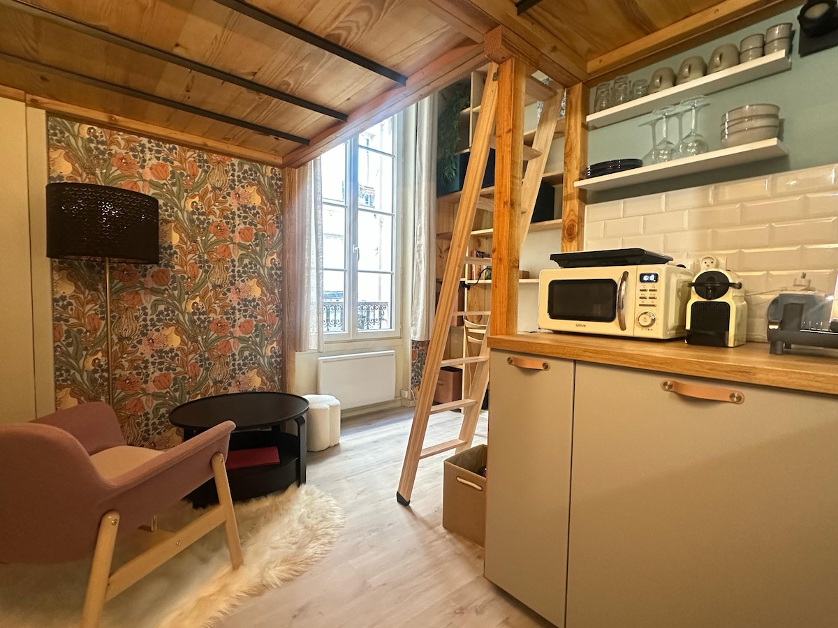 A cozy studio space features a stylish armchair beside a low coffee table. A wooden ladder leads to the mezzanine sleeping area. Natural light enters through a large window, illuminating the warm wood tones of the ceiling and furniture, while decorative wallpaper adds a pop of color.