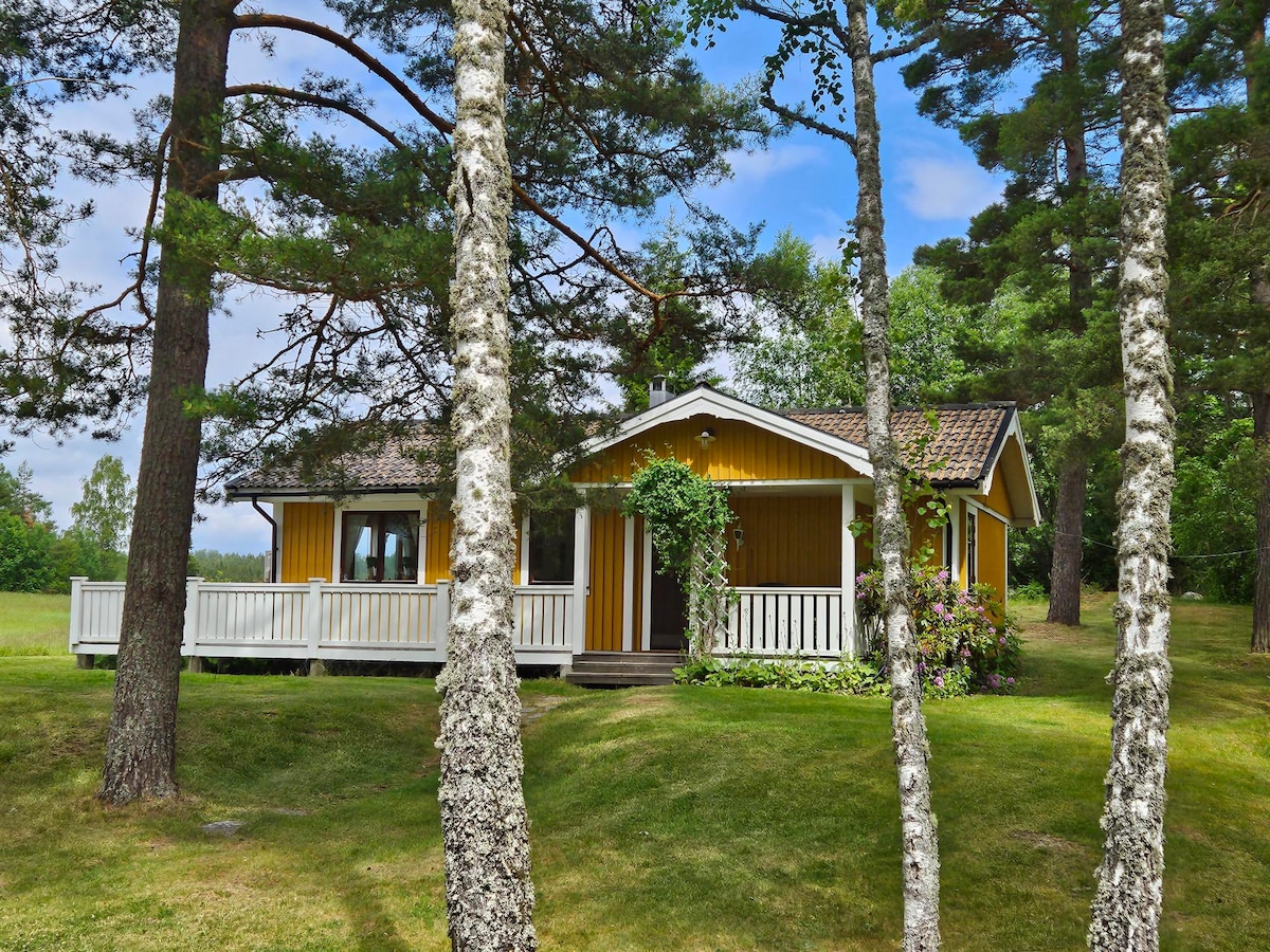 A charming cottage is framed by tall trees and lush greenery. The exterior features a yellow façade with white accents, offering a welcoming appearance. A spacious porch is visible, supplemented by colorful flowers and a neatly maintained lawn, enhancing the connection to nature.
