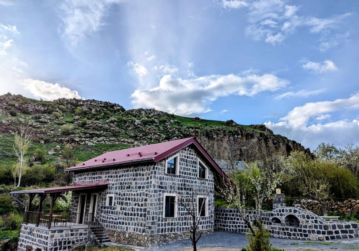 A stone house with a red roof is situated at the base of a rocky hill. Surrounding greenery adds to the scenic landscape, while a clear sky with scattered clouds enhances the tranquil setting.