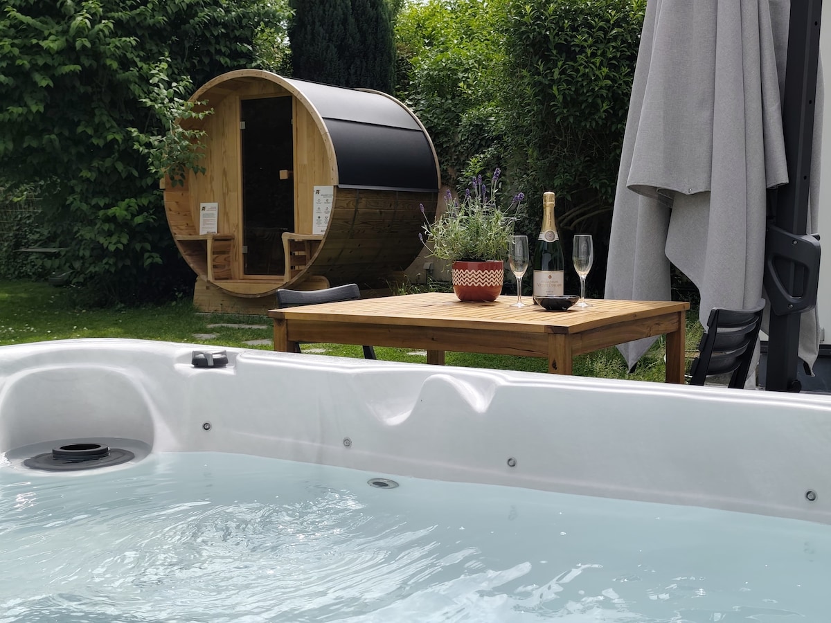 A relaxing outdoor space features a jacuzzi with bubbling water in the foreground. A wooden table is set with a bottle of champagne and two glasses, surrounded by greenery. In the background, a sauna barrel is situated under a shaded area, enhancing the tranquil ambiance.