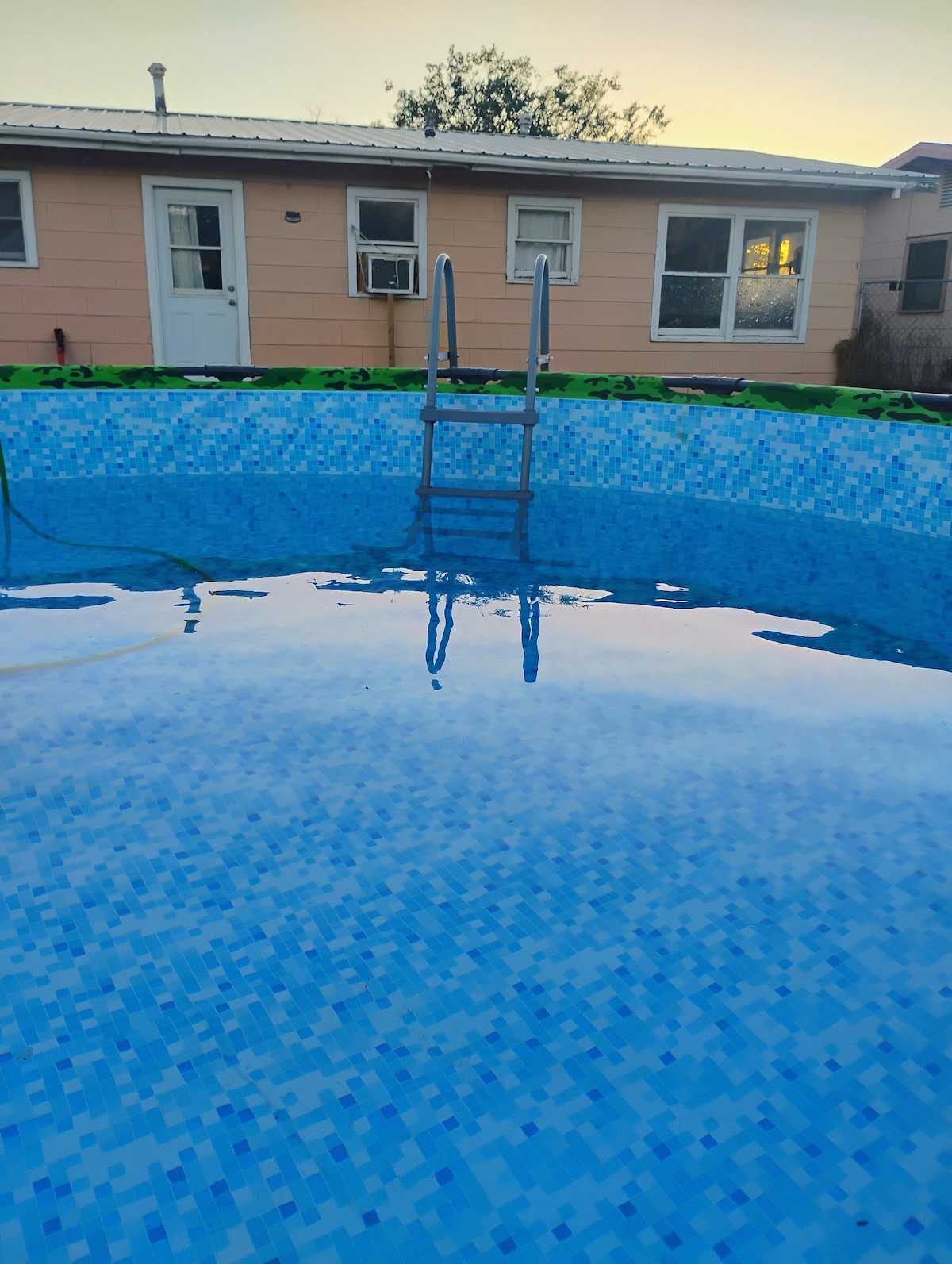 A circular swimming pool is visible, featuring a light blue tiled surface reflecting the sky. A ladder is positioned at the edge for easy access. The background shows a single-story building with multiple windows and a white door.