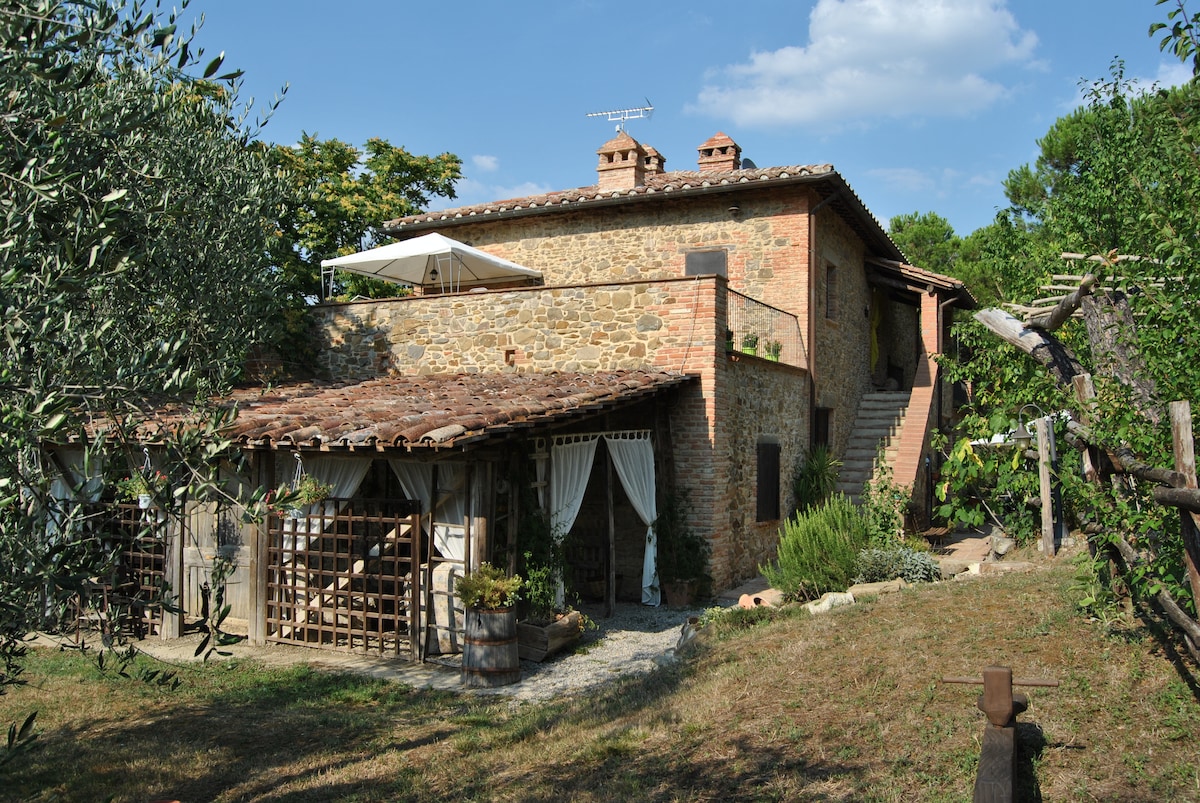 A rustic stone farmhouse is set against a clear sky. The building features a terracotta roof and large windows. Surrounding greenery includes olive trees, while a shaded patio area with draped curtains offers a relaxing outdoor space. Stairs lead to an upper level.