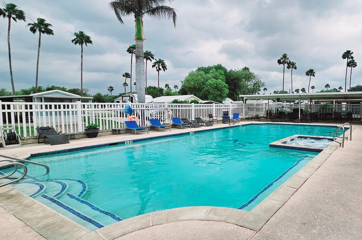 A sparkling outdoor pool is surrounded by lounge chairs, inviting relaxation. Towering palm trees frame the scene, while a hot tub is positioned nearby. The clear blue water reflects the cloudy sky, creating a tranquil setting for guests to enjoy.