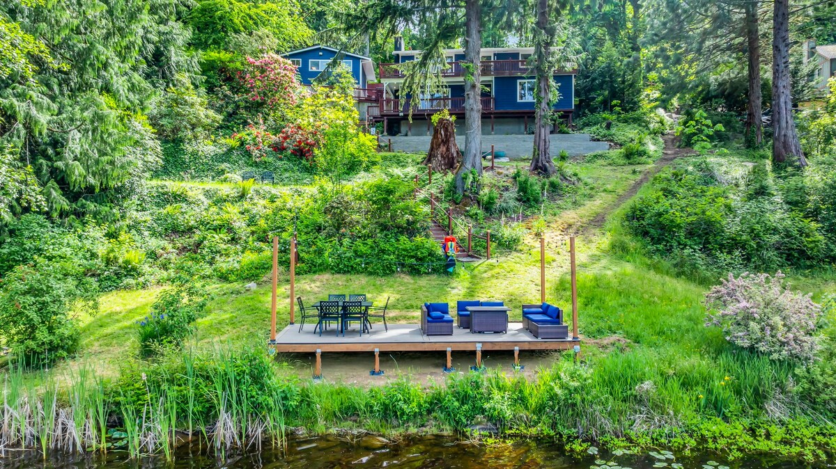A spacious dock area is shown at the water's edge, featuring a seating arrangement with blue chairs and a table. Lush greenery surrounds the area, providing a natural setting. In the background, a private unit is visible, nestled amongst the trees and hillside.
