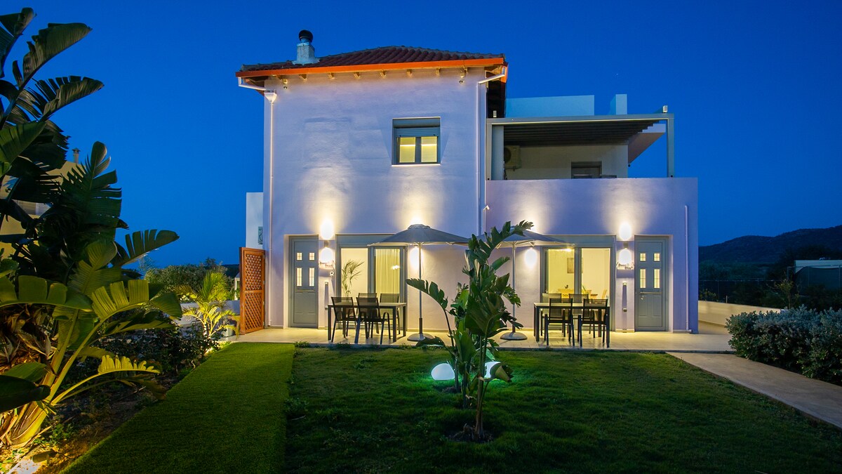 The exterior of the modern apartment is illuminated by warm lighting, revealing a spacious garden area with green grass. Tables and chairs are set under a covered terrace, providing a comfortable outdoor dining space. The structure features a light pastel color and a simple roof design.