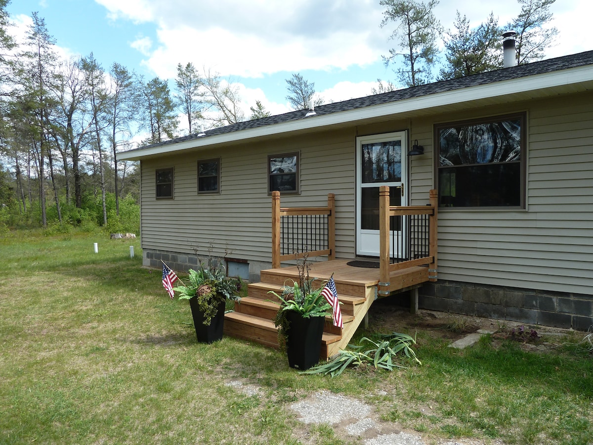 The entrance to the cabin is highlighted by a wooden deck featuring a set of steps leading to a glass door. Potted plants with small American flags are positioned on either side of the entryway. The surrounding area includes a grassy expanse and scattered trees in the background.