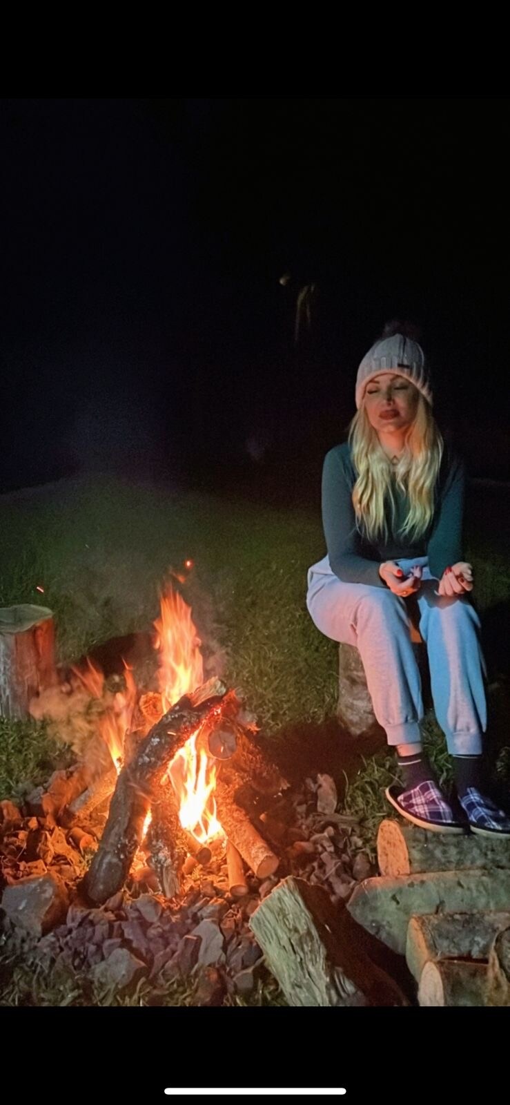 A warm campfire crackles amidst a dark night, surrounded by stones and logs. Flickering flames illuminate the scene, reflecting on a person seated on a log, dressed in relaxed attire. The natural surroundings suggest a peaceful outdoor setting, ideal for gathering and contemplation.