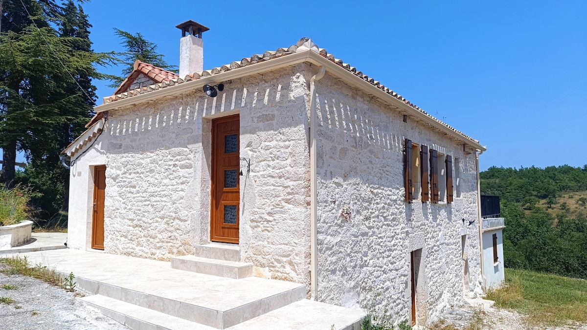 A charming stone house is featured, showcasing a classic architectural style with a tiled roof and wooden shutters. The exterior is set against a clear blue sky, surrounded by greenery, offering a peaceful atmosphere. A patio area is visible, accessible from the entrance.