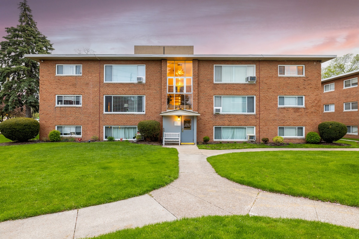 A brick apartment building is framed by well-maintained green grass and shrubs. A clean pathway curves toward the entrance, which is flanked by large windows. Warm light glows from within, highlighting the inviting atmosphere as dusk approaches.