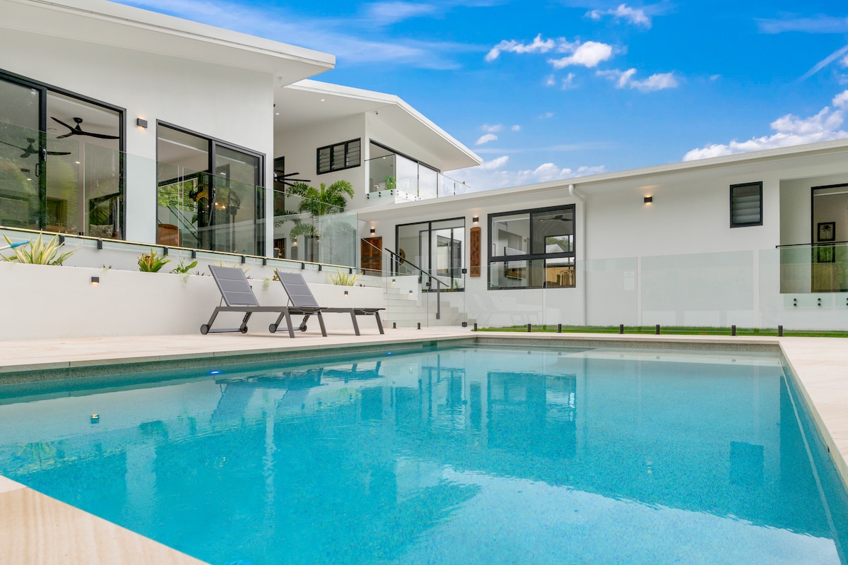 A serene pool area features a crystal-clear swimming pool embraced by modern seating. The surrounding landscape includes fresh greenery, enhancing the outdoor space. Two loungers are positioned beside the pool, creating an inviting atmosphere for relaxation under clear blue skies.