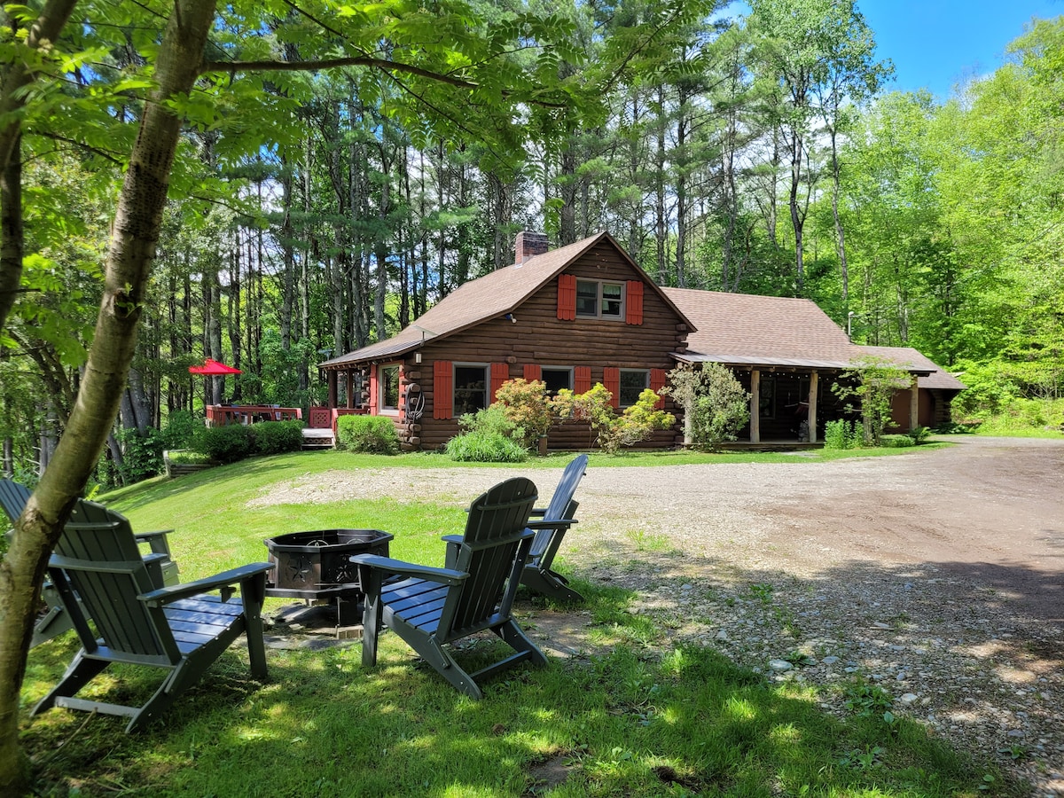 A rustic country cabin is surrounded by lush greenery and tall trees. A gravel driveway leads up to the cabin, which features red shutters and a welcoming porch. Four Adirondack chairs are arranged around a firepit, inviting relaxation in the outdoor space.
