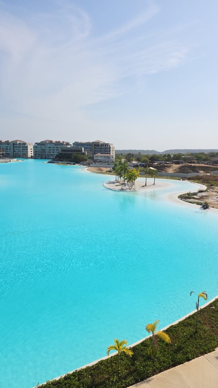 A wide view of the Crystal Lagoon features vibrant turquoise waters surrounded by sandy shores and lush greenery. The calm surface reflects the clear blue sky, creating a serene environment ideal for relaxation and water activities.