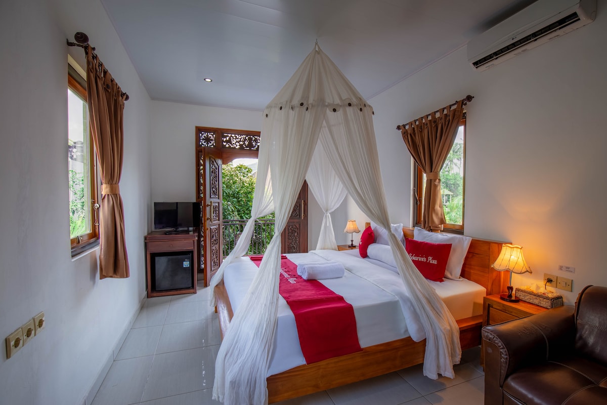 The bedroom features a spacious bed draped with a white canopy, complemented by red decorative pillows. Natural light filters through the windows, revealing a lush outdoor view. A wooden cabinet and a flat-screen TV add functionality, while warm lamps create a soft glow.