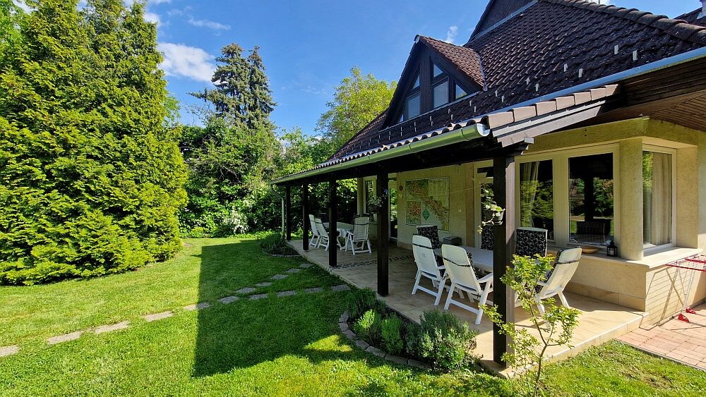 A spacious covered terrace is seen adjacent to a well-maintained garden, surrounded by lush greenery. White lounge chairs are arranged for relaxation under the shade, inviting guests to enjoy the outdoor environment. The home features a sloped roof and a glimpse of the garden through large windows.