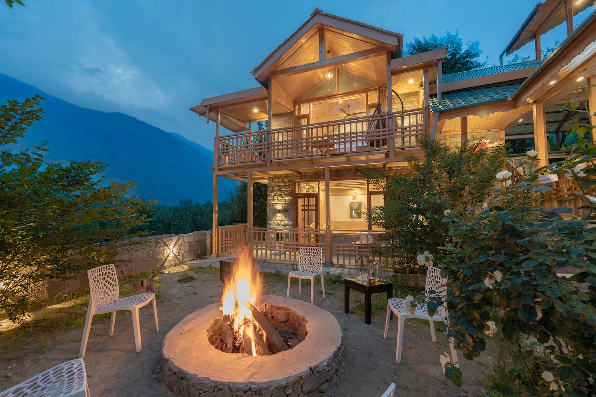 An outdoor seating area is set around a stone fire pit, surrounded by white chairs. The villa's wooden architecture is visible, showcasing multiple balconies and large windows. The setting is enhanced by the soft glow of lights against the backdrop of mountains and an evening sky.