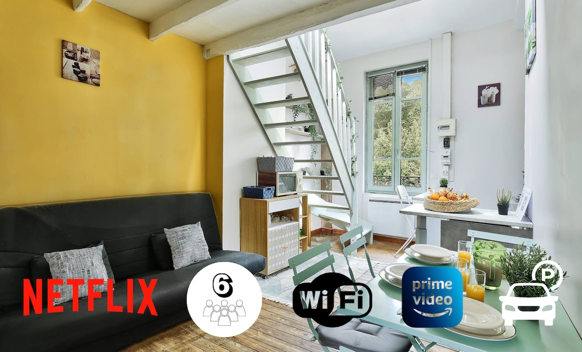 A bright living space features a dark sofa and a light green dining area. Natural light enters through the large window, illuminating the stylish decor. A staircase leads to the mezzanine level, while a dining table is set for six, creating an inviting atmosphere.