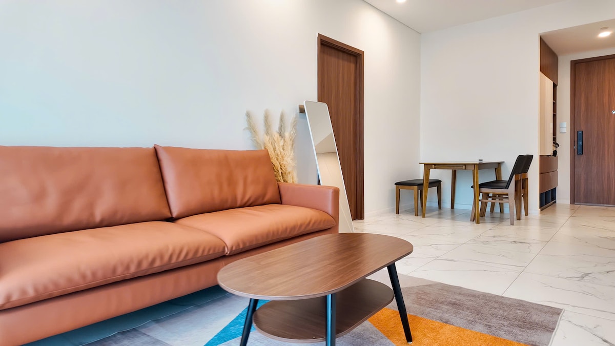 A modern living area features a light brown leather sofa positioned across from a low coffee table. A full-length mirror reflects soft light, while a dining space with a wooden table and black chairs is visible in the background. Neutral-coloured walls enhance the airy feel.