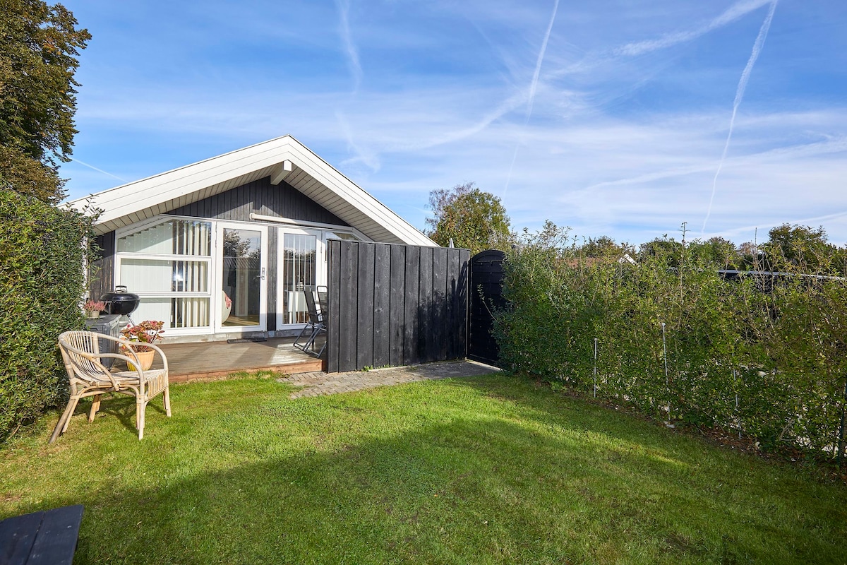 The exterior of the guest cabin features a well-maintained garden area with green grass and hedges. A patio is visible, furnished with a small table and chairs. The cabin's entrance is framed by large glass doors that invite natural light into the space.