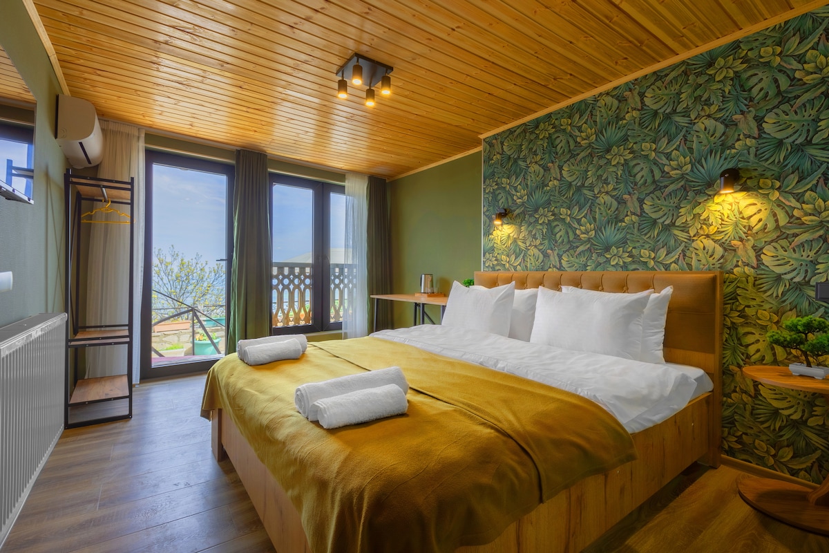 A spacious bedroom is highlighted by a wooden ceiling and botanical wallpaper. A large bed is dressed in soft linens with neatly folded towels on top. Floor-to-ceiling windows offer natural light and views of the outdoor area, enhancing the cozy ambiance of the room.