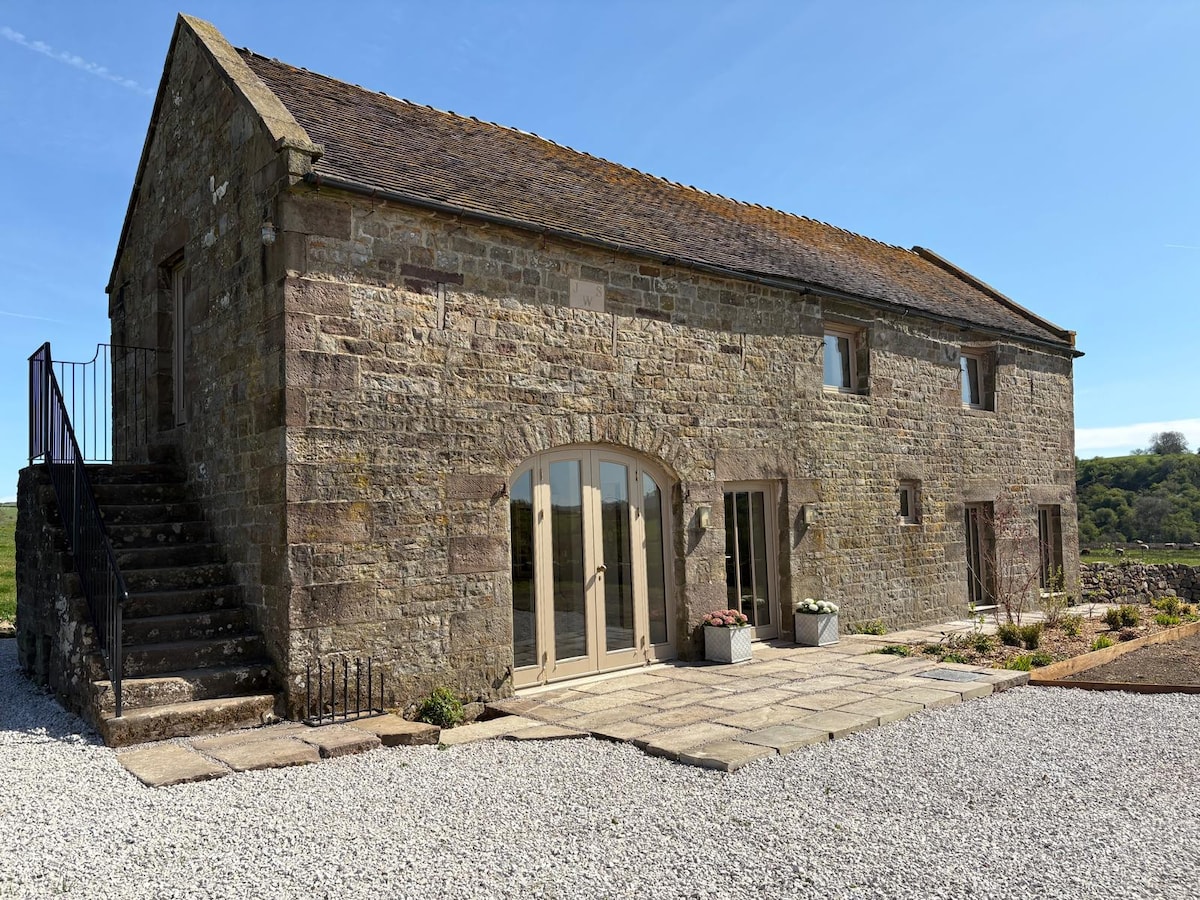 The exterior of a charming stone barn is shown, featuring large glass doors that offer a view into the interior. A set of stone steps leads to the entrance, surrounded by a gravel pathway and landscaped with potted plants, all set against a clear blue sky.