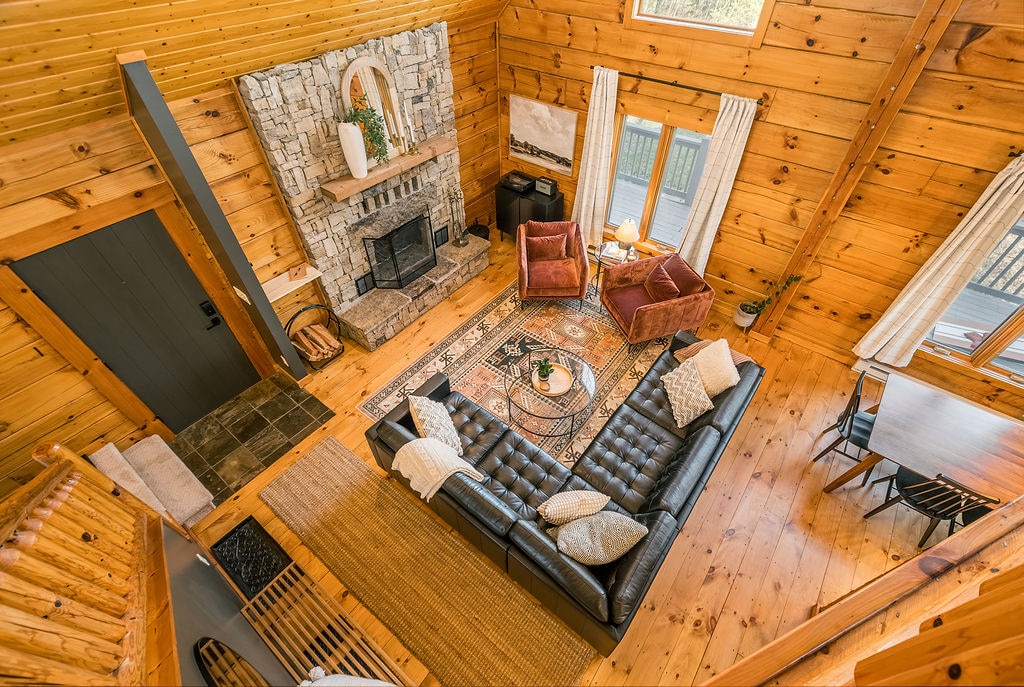 A spacious living room features a wood-burning fireplace set in a stone surround, complemented by two cozy armchairs. A dark leather sectional encircles a round coffee table on a patterned rug. Large windows let in natural light, with wooden walls enhancing the cabin's charm.