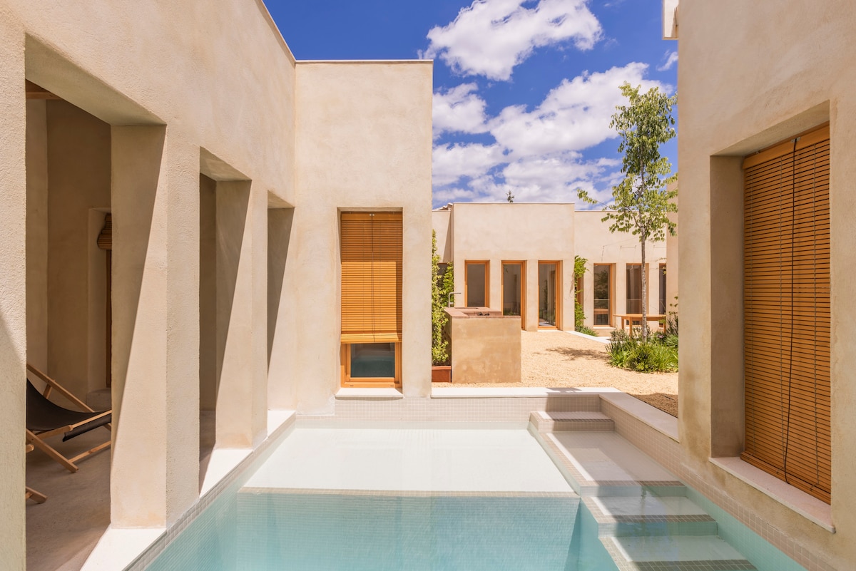 An interior courtyard features a serene pool surrounded by light-colored walls. Sunlight filters through wooden slatted shades on the windows. Lounge chairs are positioned near the pool, creating a calm space for relaxation amidst the greenery and natural elements.