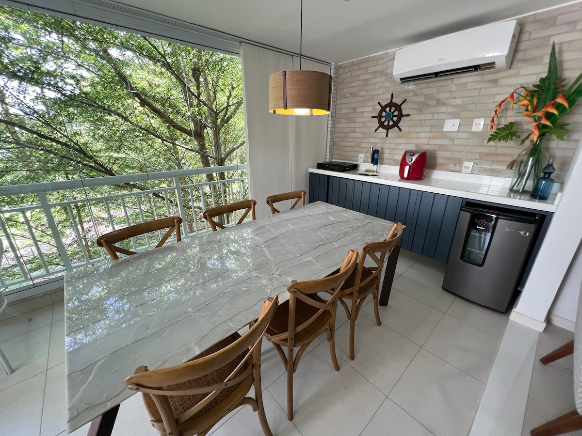 A dining area is highlighted by a large rectangular table surrounded by six wooden chairs. Natural light filters through large windows, showcasing views of greenery. A small kitchenette features essential appliances, including a coffee maker and a refrigerator, set against a subtly textured wall.