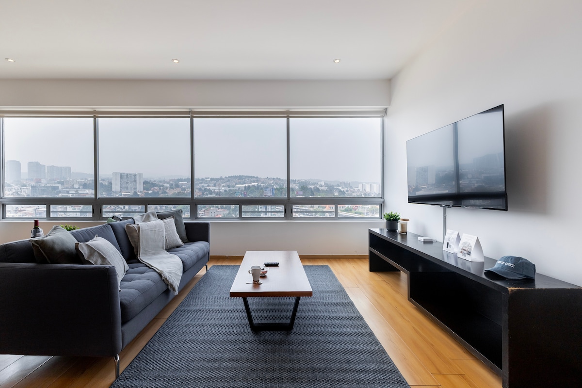 A spacious living area displays large windows that frame city views. A dark sofa with cushions is positioned next to a wooden coffee table, complemented by a textured area rug. A media console holds a flat-screen television, enhancing the modern and inviting design.