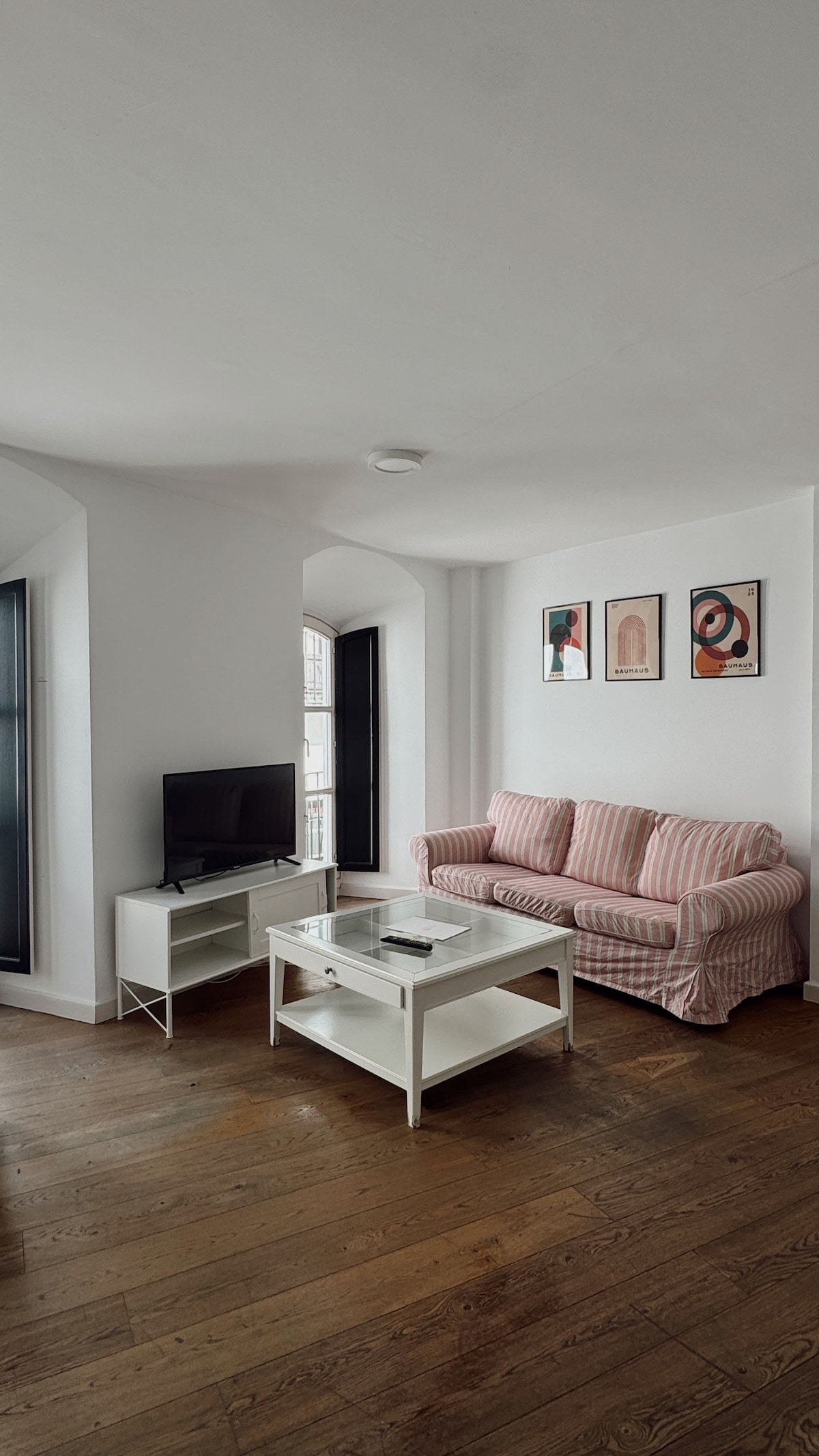 A cozy living area is presented with a striped sofa and a coffee table. A television sits on a stand, and two tall windows allow natural light to illuminate the wooden flooring. Artwork adorns the walls, adding character to the space.