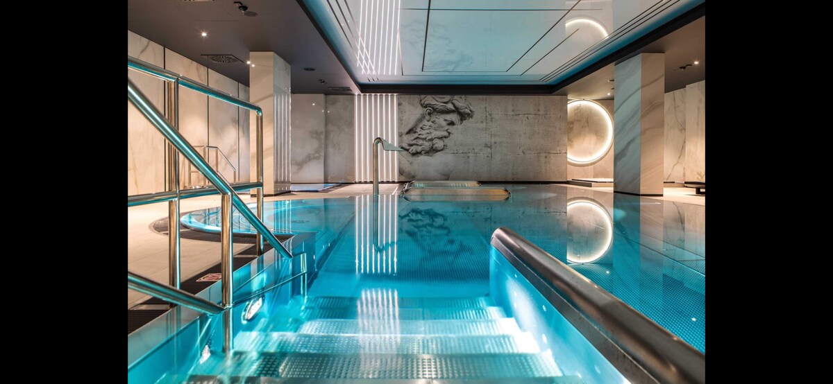 An indoor pool area is illuminated by soft lighting, showcasing clear blue water. Elegant steps lead into the pool, with marble walls displaying a sculptural relief. The tranquil environment is complemented by modern design elements, creating a serene space for relaxation.