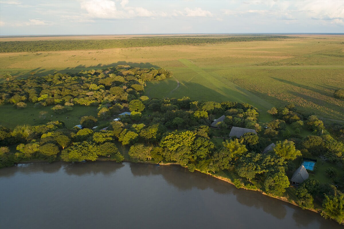 An expansive landscape is captured from above, showcasing lush greenery bordering a calm river. The open fields extend into the distance, complemented by scattered trees and a few structures nestled near the water's edge. The tranquility of the natural environment is evident.